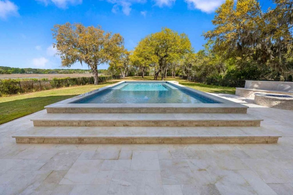 Modern rectangular swimming pool with elevated design, stone patio, built-in seating area, and landscaped backyard view